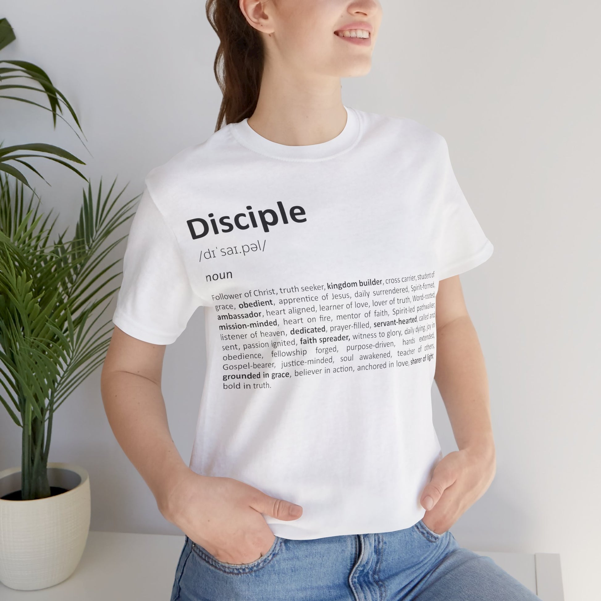 Woman wearing a white t-shirt with 'Disciple' definition text, standing indoors next to a plant.