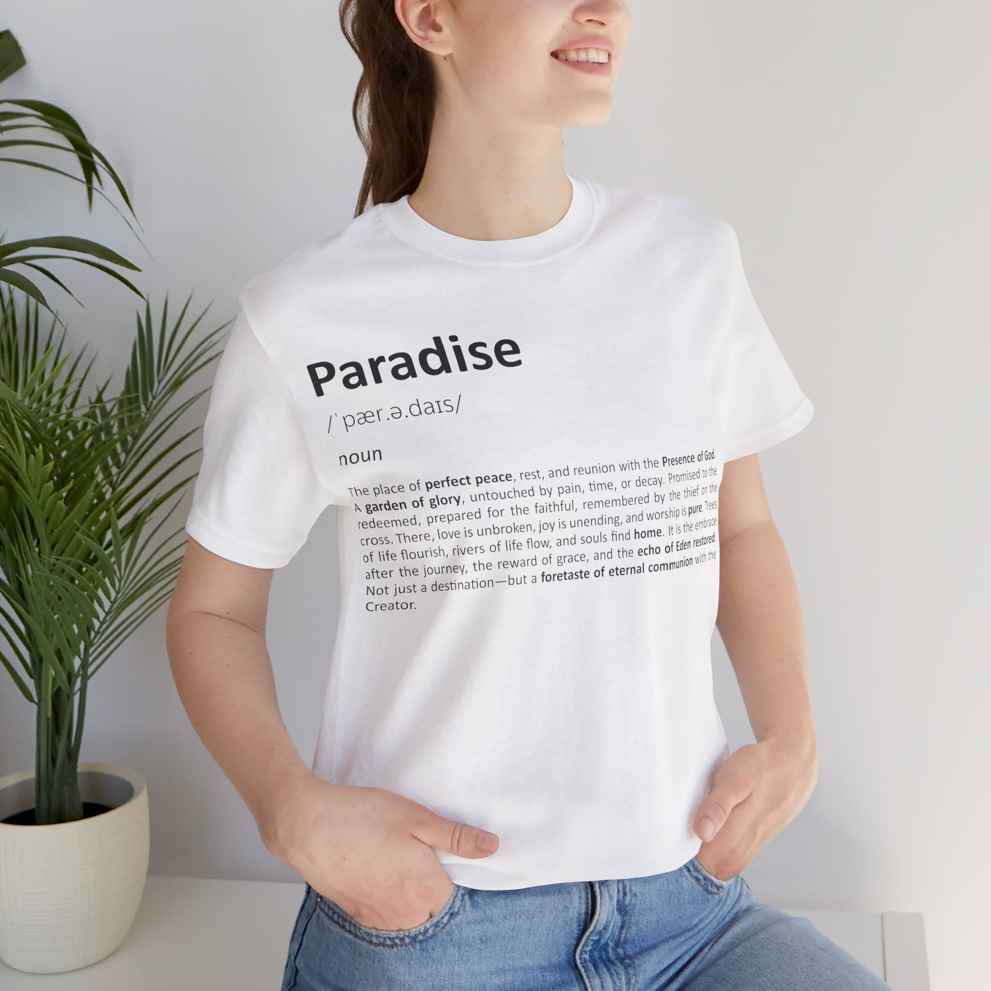 Person wearing a white t-shirt with 'Paradise' definition text, standing indoors next to a plant.