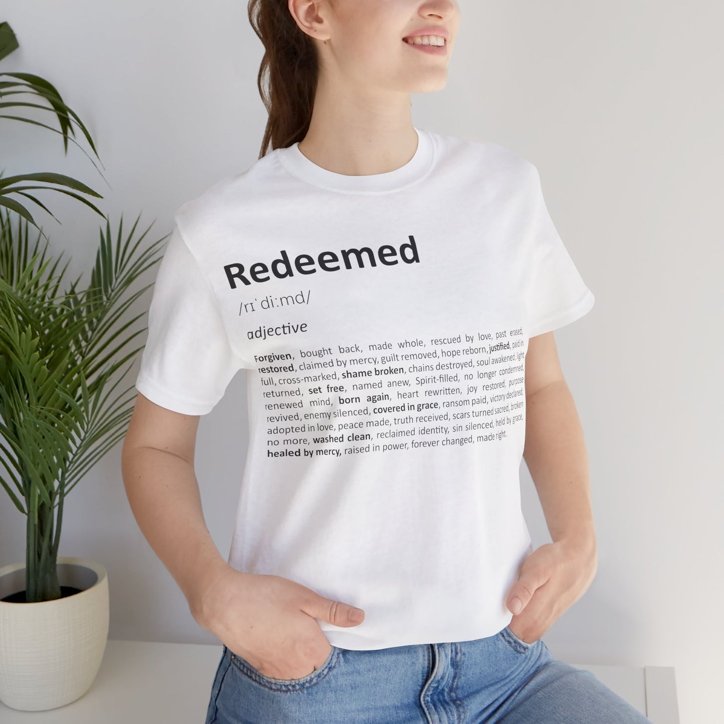 Person wearing a white t-shirt with 'Redeemed' definition text, standing indoors next to a plant.